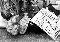 A man lies on the ground next to his wheelchair, holding a sign that says 'Nursing Homes Kill.'